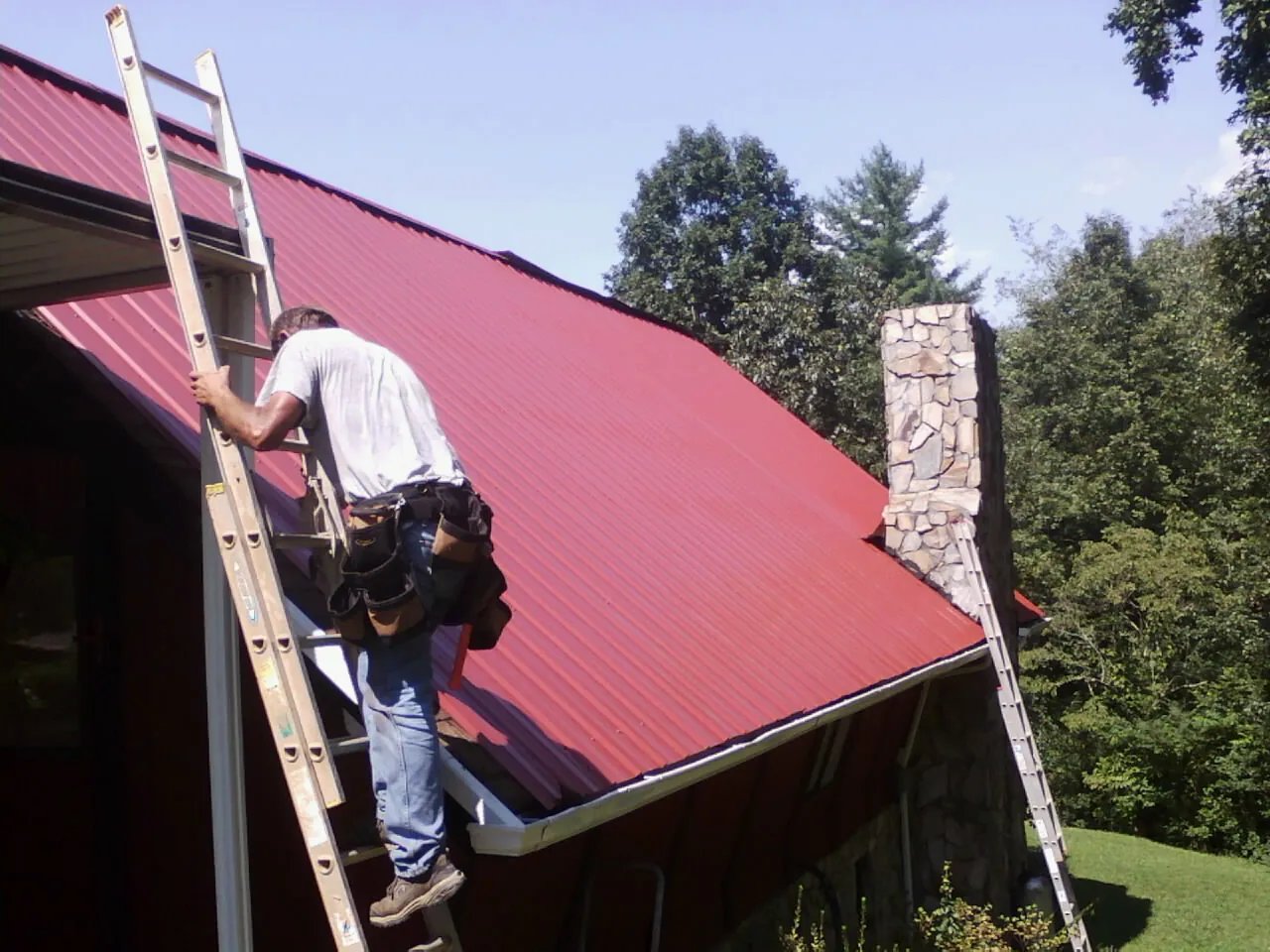 Roofer on a ladder beside a red metal roof and stone chimney — Soffit & Fascia Repair by Roofing Brothers in Glide